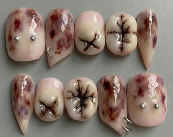 Puede incluir: Un conjunto de diez uñas postizas con un diseño único. Las uñas presentan una base rosa pálido con patrones moteados granate y marrón. Algunas uñas tienen forma de dientes, con diseños detallados de raíz y esmalte. Se añaden tachuelas y anillos plateados.