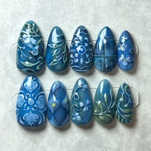 May include: A set of ten artificial nails with a blue and white floral and geometric design. The nails are oval-shaped and feature intricate patterns, including leaves, flowers, and abstract shapes. The color palette includes shades of blue and white.