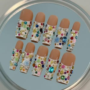 May include: A collection of ten artificial nails featuring a nude base and white tips, decorated with an assortment of colourful gems, pearls, and gold details. The decorations include stars, moons, and various shapes, creating a glamorous and striking design.