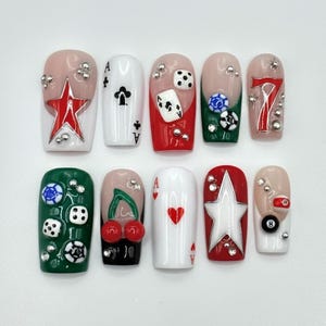 May include: A set of ten artificial nails with a casino theme. Designs include playing cards, dice, stars, and the number seven. The nails feature red, green, white, and nude colours, with silver embellishments.