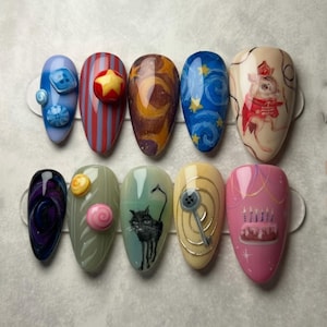 May include: A collection of ten almond-shaped, painted fingernails. Designs include a blue nail with buttons, a red and purple striped nail with a star, and a nail with a black cat illustration. Other nails feature abstract art and a birthday cake.