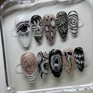 May include: A set of ten press-on nails with unique, hand-painted designs. The nails feature a variety of black and white illustrations, including eyes, faces, and abstract patterns, on a white or nude base. The nails are displayed in a clear plastic case.