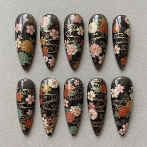 May include: A set of ten almond-shaped artificial nails. Each nail has a black, glittery base and is decorated with floral patterns in pink, white, orange, and gold, with gold wave accents.