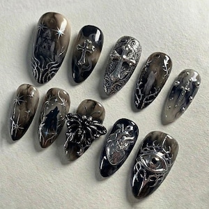 May include: A set of ten long, oval-shaped press-on nails with a dark, gothic aesthetic. Each nail features unique designs, including crosses, a wolf, a butterfly, and an eye, all in silver or black. The nails have a marbled effect.