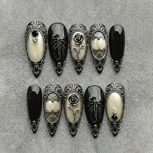 May include: A set of ten long, pointed, black and silver press-on nails. Some nails feature ornate silver frames with cream-colored inserts, a rose, or a spider design. The nails are arranged on a gray surface.