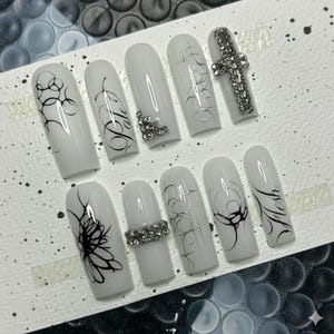 May include: A set of white press-on nails with various designs. Some nails feature black abstract swirls, silver line art, or rhinestone accents. One nail has a rhinestone cross, and another reads "flesh" in black script.