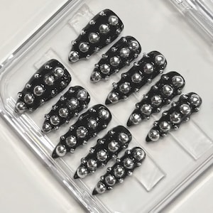 Peut inclure: Ongles noirs à presser avec des clous métalliques argentés disposés en rangées. Les ongles sont en forme de cercueil et présentés dans un récipient en plastique transparent. Un ensemble de faux ongles.