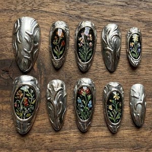 Könnte beinhalten: Eine Sammlung von zehn silberfarbenen Nagelkunststücken. Jedes Stück zeigt ein ovales Design mit einem Blumenarrangement vor dunklem Hintergrund. Die silbernen Teile haben ein geprägtes, organisches Muster.