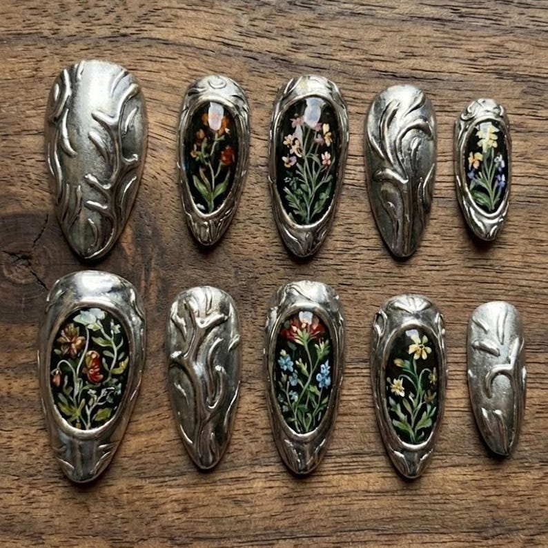 May include: A collection of ten silver-toned nail art pieces. Each piece features an oval-shaped design with a floral arrangement set against a dark background. The silver pieces have an embossed, organic pattern.