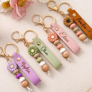 May include: Four personalized keychains featuring leather-like straps in purple, sage green, pink, and tan. Each strap is engraved with names Sophia, Olive, Elina, and Amelia. They include gold-tone lobster clasps, a flower charm, and wooden and silicone beads.