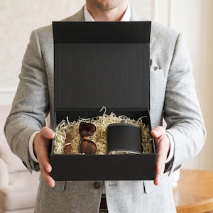 May include: A black rectangular gift box containing a black metal flask, brown-rimmed sunglasses, and a small folding tool, all nestled in light-colored shredded paper filler. The box is open with a vertical lid, presenting the items clearly.
