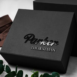 Könnte beinhalten: Eine quadratische schwarze Geschenkbox mit strukturierter Oberfläche. Auf dem Deckel befindet sich der Name Parker in einer großen, glänzend schwarzen Schreibschrift, darüber der Text THEBESTMAN in kleinerer, serifenloser Schrift.