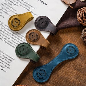 May include: Five leather magnetic clips in mustard yellow, dark brown, tan, forest green, and blue. Each clip features a circular embossed design with a laurel wreath surrounding a single letter: A, T, U, S, or K. The clips are arranged on a white page.