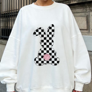 May include: White sweatshirt featuring a black and white checkered bunny graphic. The bunny has a pink floral tail. The sweatshirt is a casual, comfortable style, suitable for everyday wear. The design is centered on the front.