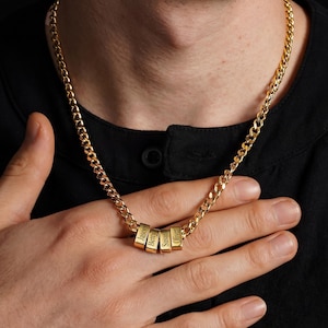 Puede incluir: Collar de cadena de oro con tres colgantes rectangulares. Cada colgante tiene un nombre grabado en una fuente serif: Jason, Betty, Susan y Oliver. El collar se lleva sobre una camisa negra.