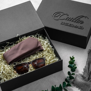 May include: A black rectangular gift box containing a folded mauve fabric accessory and dark-rimmed sunglasses nestled in gold-colored paper shred. The box lid features the embossed text Caitlin BRIDESMAID in a black script and block font.