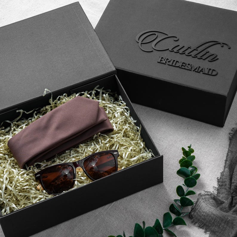 May include: A black rectangular gift box containing a folded mauve fabric accessory and dark-rimmed sunglasses nestled in gold-colored paper shred. The box lid features the embossed text Caitlin BRIDESMAID in a black script and block font.