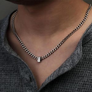 Puede incluir: Un collar de cadena de plata con un pequeño colgante rectangular. El colgante tiene la palabra "Lucas" grabada. El collar se lleva sobre una camisa gris de punto de gofre.