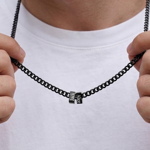 May include: Black chain necklace with two small, rectangular pendants. Each pendant has a name engraved on it: "Jack" and "Alice". The necklace is held up by a person wearing a white t-shirt.