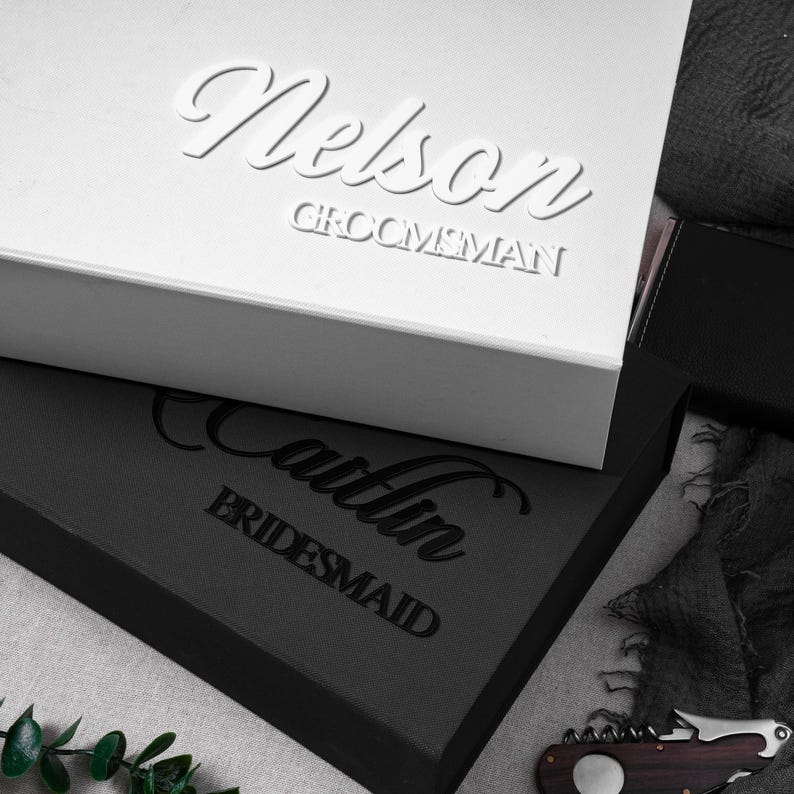 May include: A white rectangular gift box with the name Nelson and the word GROOMSMAN in raised white lettering, stacked on a black rectangular gift box with the name Caitlin and the word BRIDESMAID in raised black lettering.