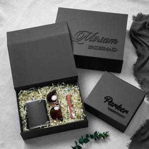 May include: Three black gift boxes with magnetic closures. One open box contains a black flask, brown sunglasses, and a bottle opener nestled in gold shredded paper. The closed boxes feature the names Miriam and Parker with titles in black.