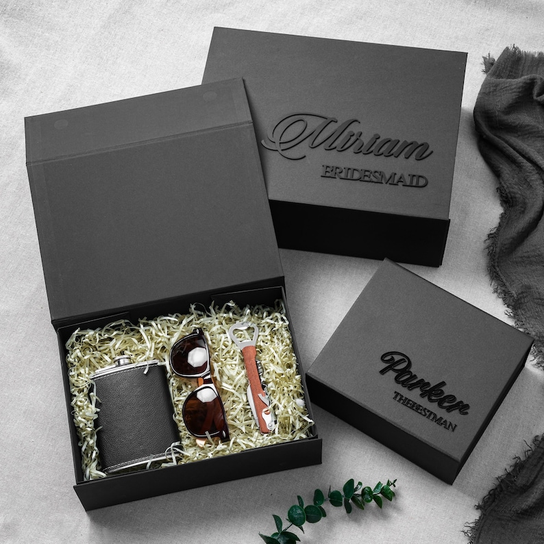 May include: Three black gift boxes with magnetic closures. One open box contains a black flask, brown sunglasses, and a bottle opener nestled in gold shredded paper. The closed boxes feature the names Miriam and Parker with titles in black.