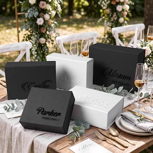 May include: A collection of rectangular gift boxes in matte black and white finishes. Each box features custom names and titles in raised lettering, including Parker The Best Man, Miriam Bridesmaid, and Prince, displayed on a wooden table setting.