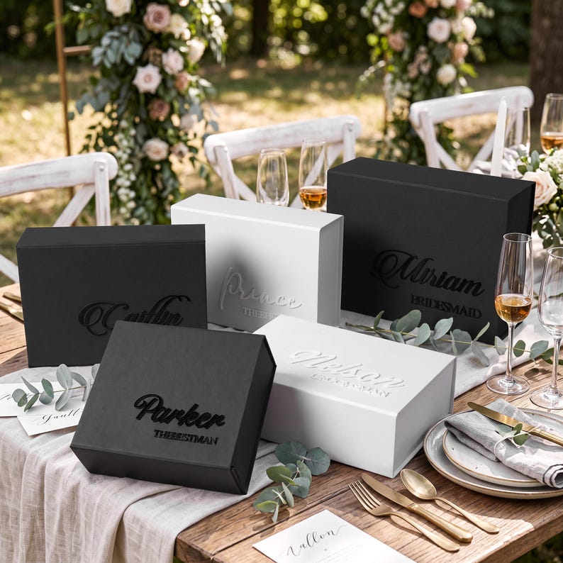 May include: A collection of rectangular gift boxes in matte black and white finishes. Each box features custom names and titles in raised lettering, including Parker The Best Man, Miriam Bridesmaid, and Prince, displayed on a wooden table setting.