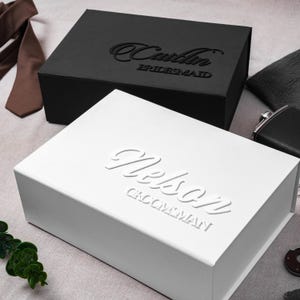 May include: A rectangular white gift box with the embossed name Nelson and the text GROOMSMAN, positioned in front of a black gift box with the embossed name Caitlin and the text BRIDESMAID, accompanied by a brown necktie and a black flask.