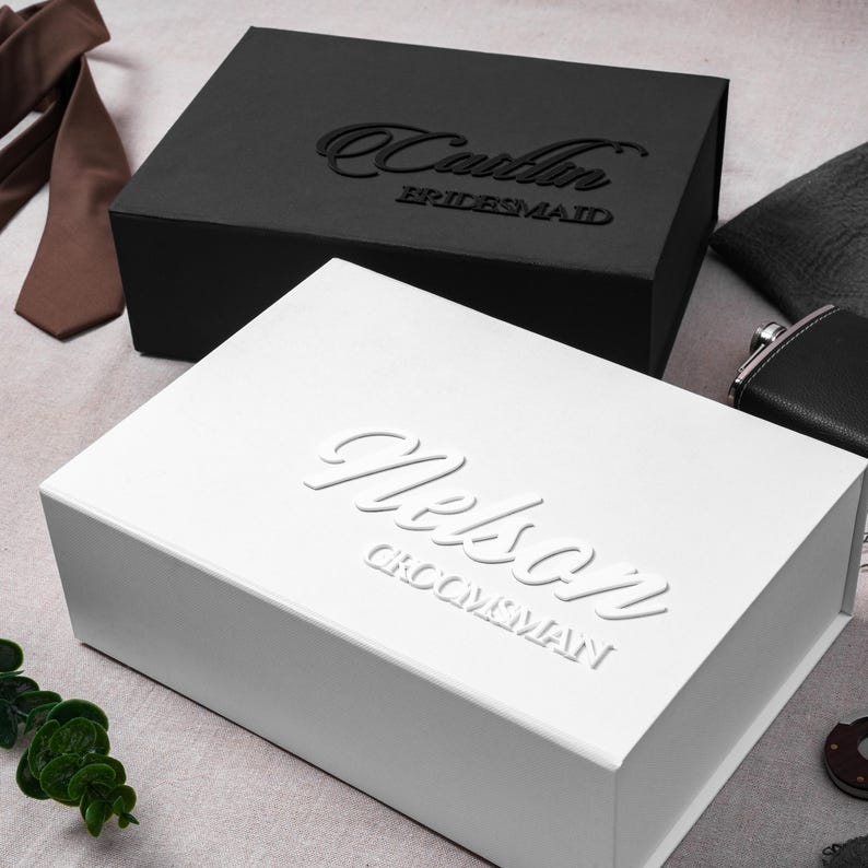 May include: A rectangular white gift box with the embossed name Nelson and the text GROOMSMAN, positioned in front of a black gift box with the embossed name Caitlin and the text BRIDESMAID, accompanied by a brown necktie and a black flask.