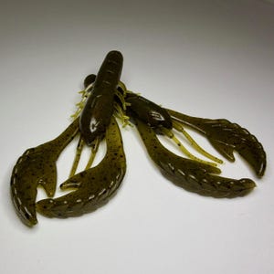 Peut inclure: Deux leurres en plastique souple vert foncé en forme d'écrevisse, avec des motifs tachetés et des accents jaunes. Les leurres sont conçus pour imiter l'apparence d'une écrevisse. Les pinces et les pattes sont déployées, et le corps est segmenté.