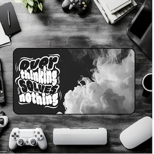 May include: Black and white desk mat with the text "Overthinking Solves Nothing." The mat features a black background with white text and a white cloud-like design. A white mouse, keyboard, and game controllers are visible.