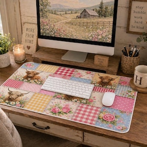 May include: A patchwork desk mat featuring Highland cows, flowers, and gingham patterns in pink, yellow, and blue. The mat has a white keyboard and mouse on top. A computer monitor and other desk accessories are in the background.