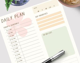 May include: A daily planner with a floral design, a pen, and a cup of coffee. The planner includes sections for scheduling, top priorities, and a to-do list. A bouquet of pink flowers sits in a vase.