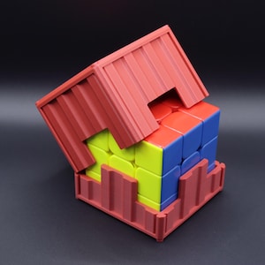 May include: A red 3D-printed container partially encases a multi-colored Rubik's Cube. The cube's faces display red, blue, and yellow squares. The container has a ridged design, and the background is black.