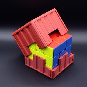 May include: A multi-colored 3x3 puzzle cube featuring yellow, blue, and red faces, partially enclosed within a red, corrugated, shipping container-style storage case that has a removable top section and a base with vertical ribbed walls.