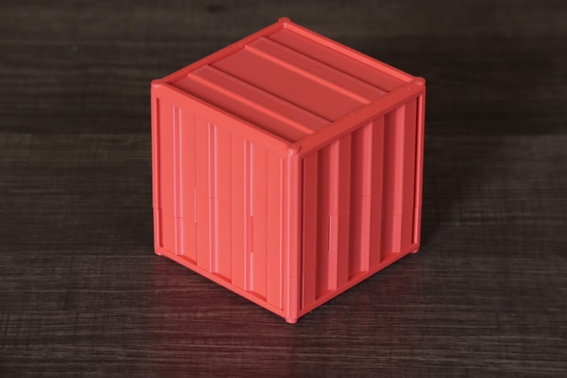 May include: A small, cube-shaped storage container in a vibrant coral-red color. The exterior features a corrugated, ribbed texture that mimics the appearance of a standard industrial shipping container, complete with vertical and horizontal lines.