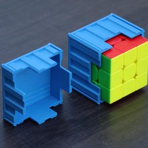 May include: A multi-colored 3x3 puzzle cube partially encased in a blue plastic container designed to resemble a shipping container. The puzzle cube features red, yellow, and green square panels on its visible faces, while a matching blue lid sits nearby.