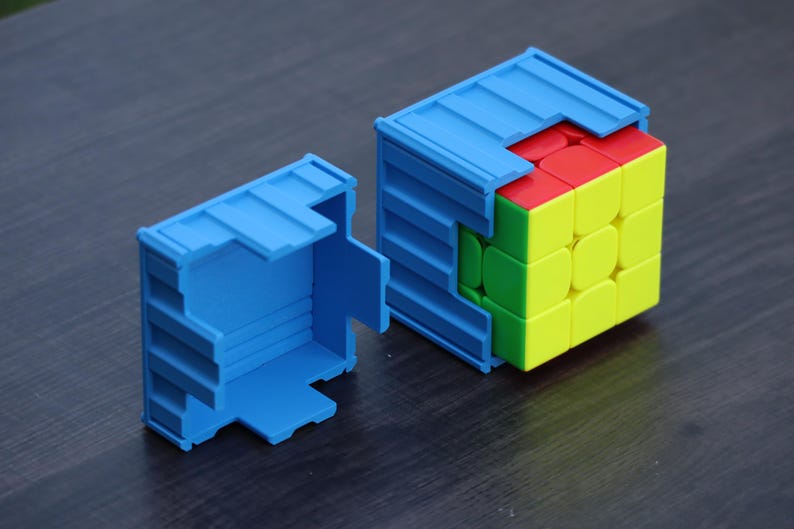 May include: A multi-colored 3x3 puzzle cube partially encased in a blue plastic container designed to resemble a shipping container. The puzzle cube features red, yellow, and green square panels on its visible faces, while a matching blue lid sits nearby.