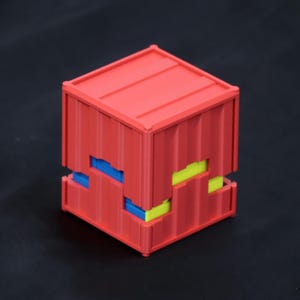 May include: A red cube-shaped container with a corrugated texture on its exterior surfaces. The structure features a horizontal gap around the middle, revealing small, alternating blue and yellow rectangular blocks nested within the red frame.