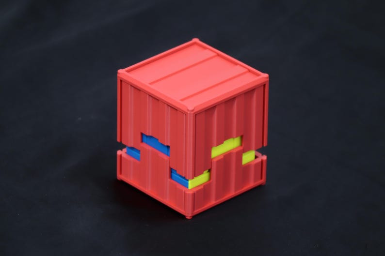 May include: A red cube-shaped container with a corrugated texture on its exterior surfaces. The structure features a horizontal gap around the middle, revealing small, alternating blue and yellow rectangular blocks nested within the red frame.