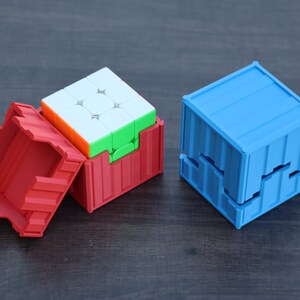 May include: A multi-colored 3x3 puzzle cube partially inside a red plastic container with a ribbed exterior design, alongside a separate blue plastic container with a matching ribbed design that is closed and shaped like a cube.