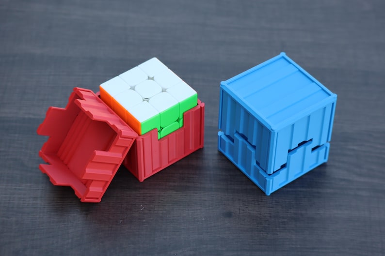 May include: A multi-colored 3x3 puzzle cube partially inside a red plastic container with a ribbed exterior design, alongside a separate blue plastic container with a matching ribbed design that is closed and shaped like a cube.