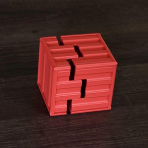 May include: A red cube-shaped puzzle box featuring a corrugated, shipping container-style texture on its exterior surfaces. The object is constructed from two interlocking pieces that form a distinct, jagged seam running across the faces.