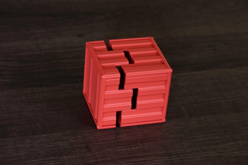May include: A red cube-shaped puzzle box featuring a corrugated, shipping container-style texture on its exterior surfaces. The object is constructed from two interlocking pieces that form a distinct, jagged seam running across the faces.