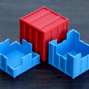 May include: A red shipping container-style box with a closed lid, positioned behind two open blue containers. Each container features vertical ribbed walls and a square base, mimicking the industrial design of modular freight storage units.