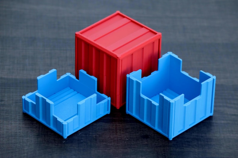 May include: A red shipping container-style box with a closed lid, positioned behind two open blue containers. Each container features vertical ribbed walls and a square base, mimicking the industrial design of modular freight storage units.
