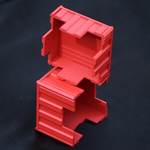 May include: A bright red, rectangular, hollow plastic container with a ribbed exterior texture. The object consists of two interlocking sections, featuring an open top and side panels with horizontal ridges, designed with a modular, box-like structure.