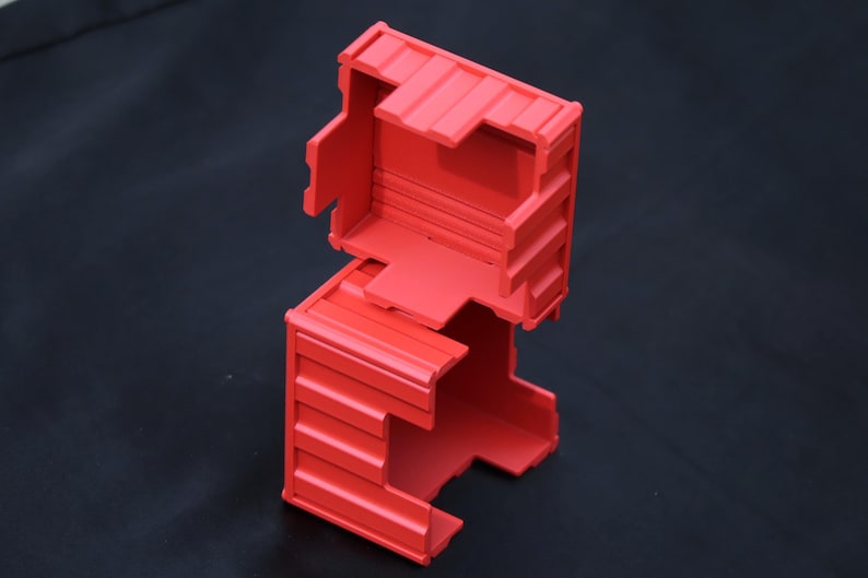 May include: A bright red, rectangular, hollow plastic container with a ribbed exterior texture. The object consists of two interlocking sections, featuring an open top and side panels with horizontal ridges, designed with a modular, box-like structure.