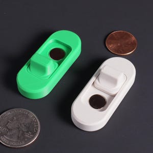 May include: Two oval-shaped plastic objects, one bright green and one white, each featuring a raised rectangular tab and a circular dark metal magnet insert. A US quarter and a copper penny are placed nearby for scale comparison.